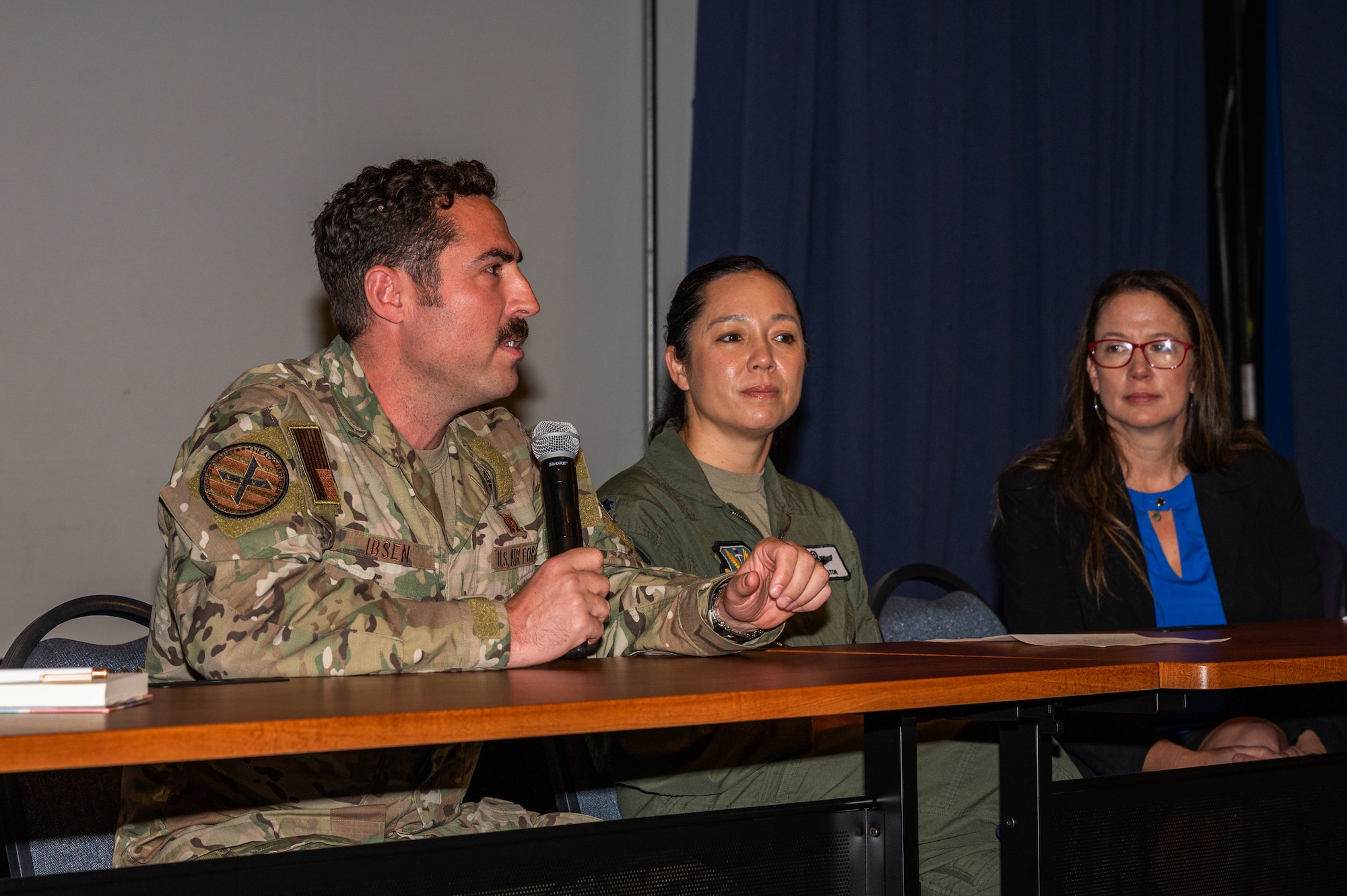 A U.S. Military members sits at a table holding a microphone and speaking.