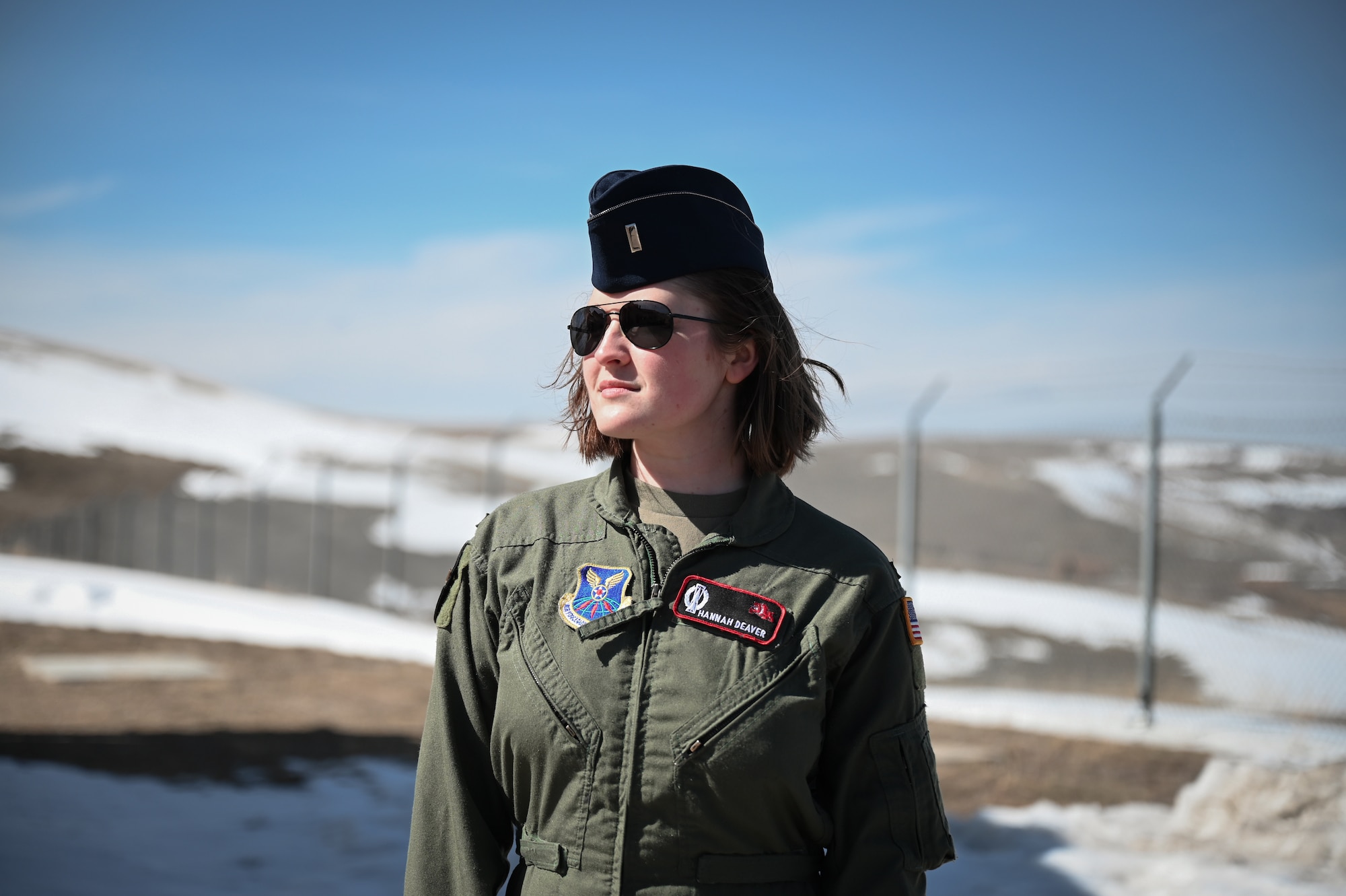 A woman in a green flight suit, blue flight cap and black sunglasses stands in the center of the photo with snow in the background.