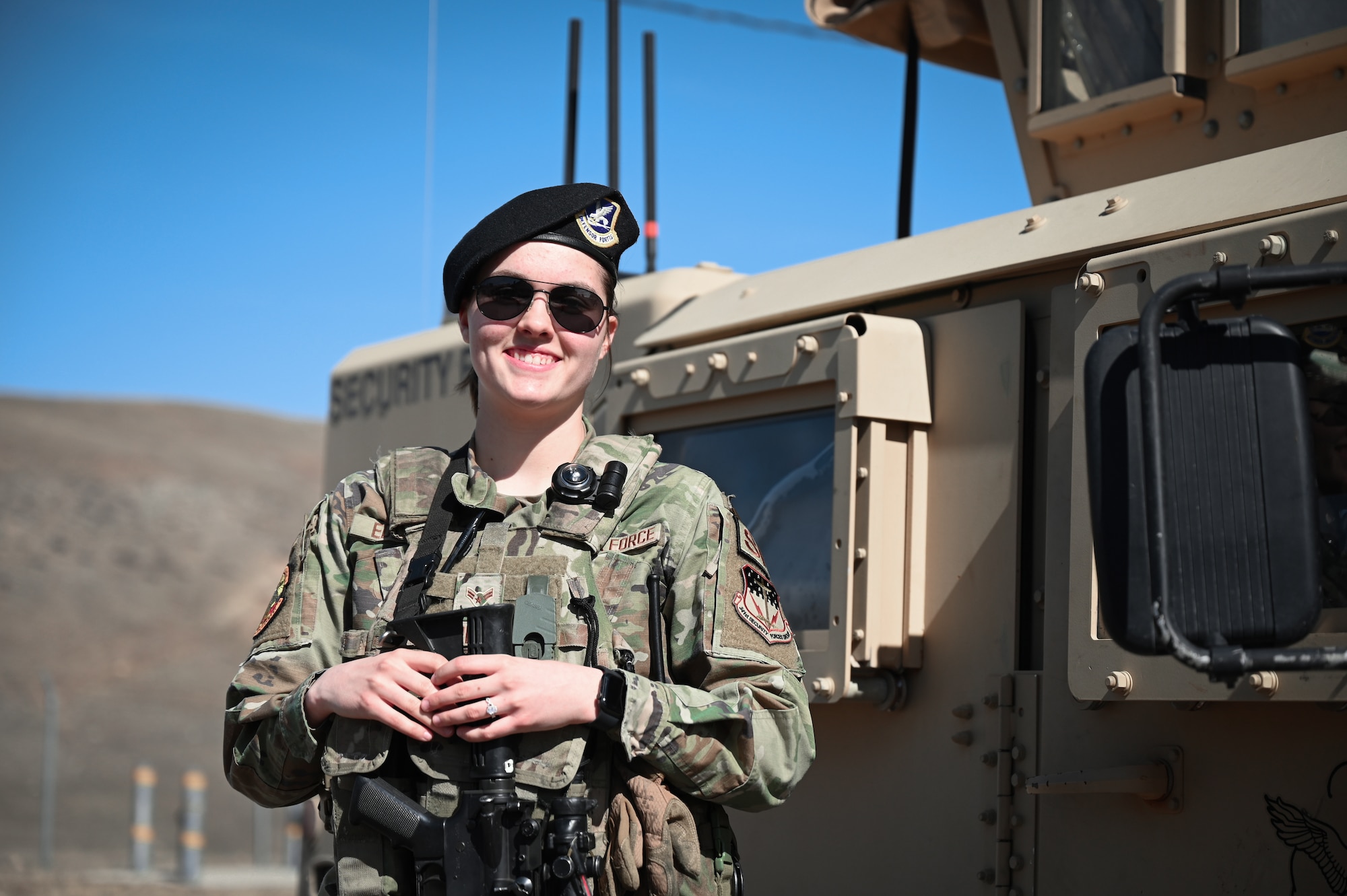A smiling woman in military uniform stands beside a military armored vehicle.