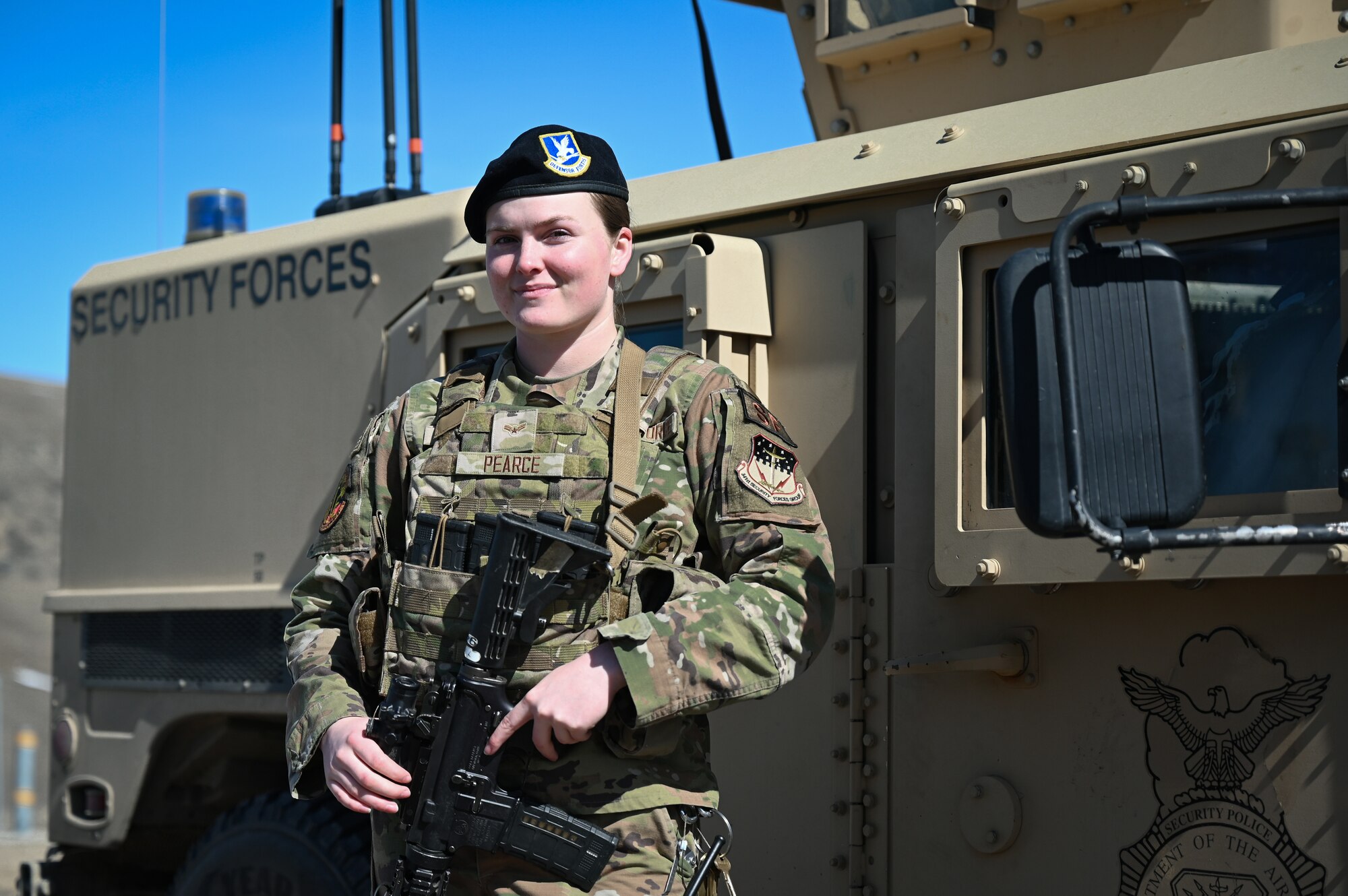 A woman in military uniform and tactical vest stands with her weapon beside a military armored vehicle.