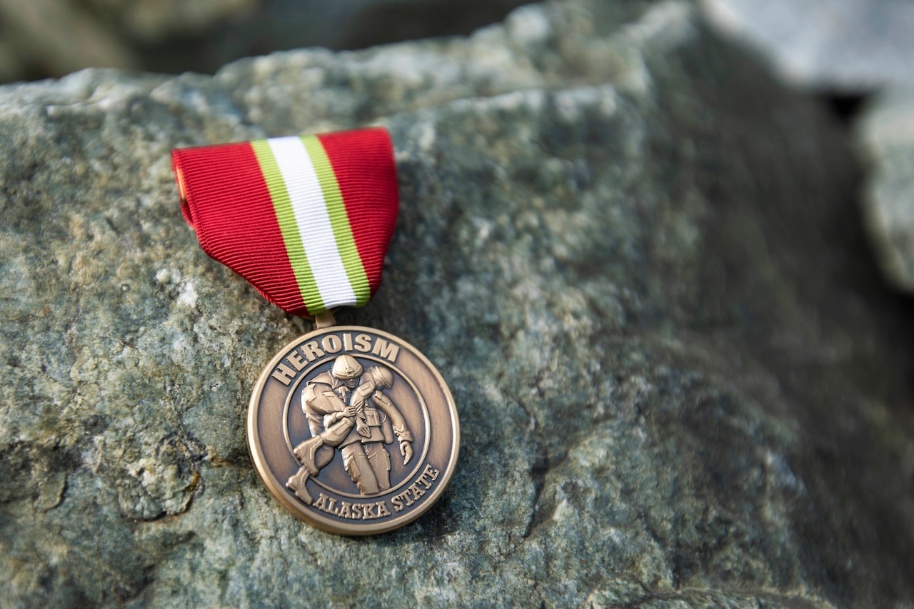 A red ribbon with center white and gold stripes and a medal that reads Heroism Alaska State with a service member carrying another in the center.