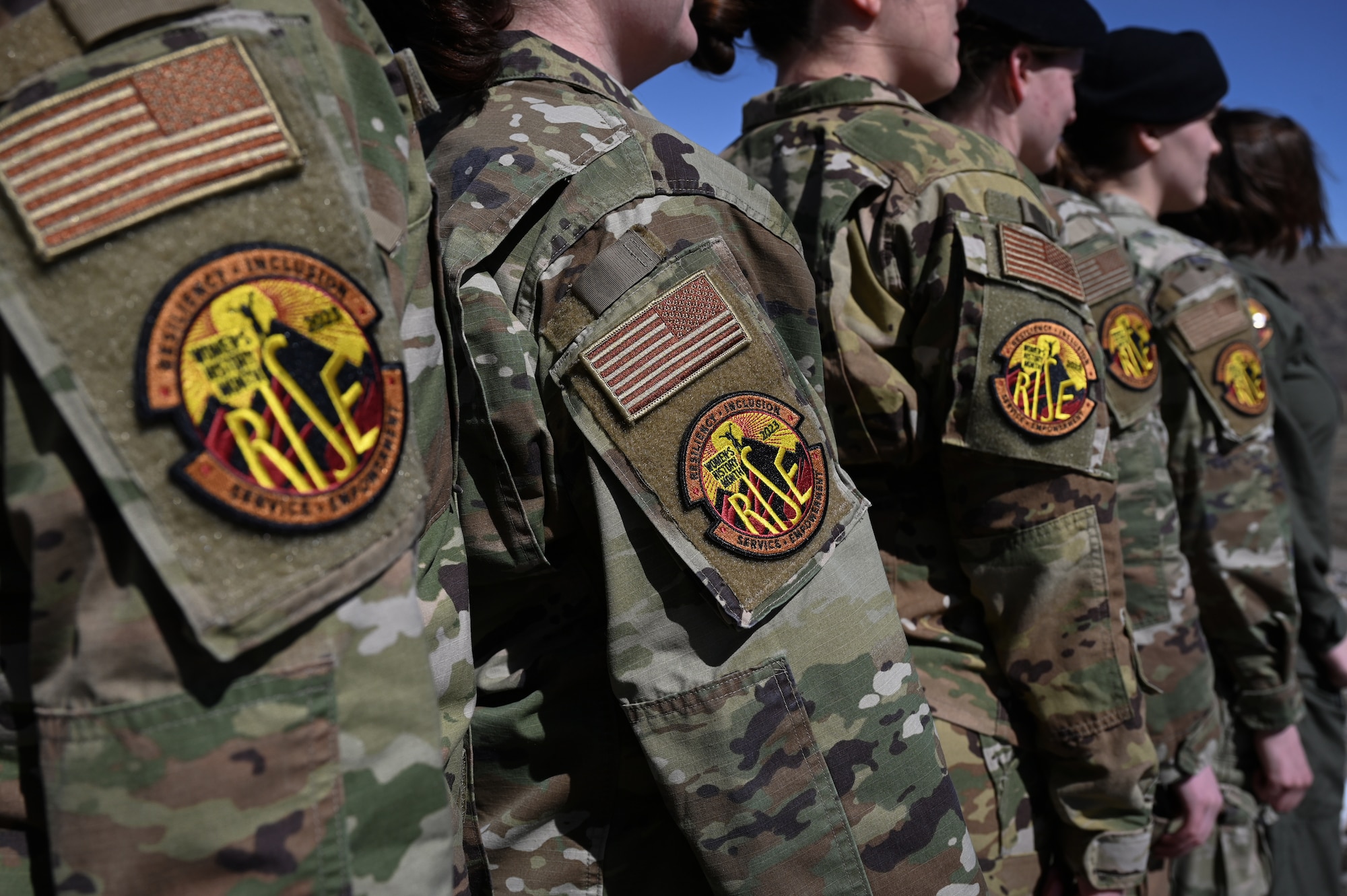 The sleeves of military uniforms are displayed in a row and adorned with a yellow and orange patch which reads, "RISE."