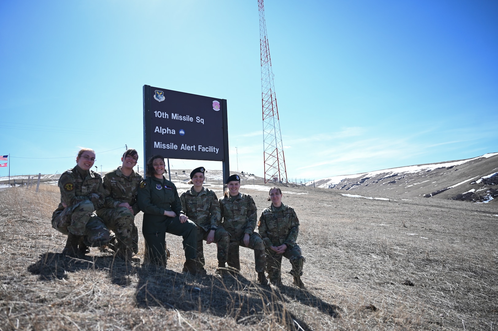 Six women in military uniform kneel in front of a sign that reads "10th Missile Sq/ Alpha 01/ Missile Alert Facility."