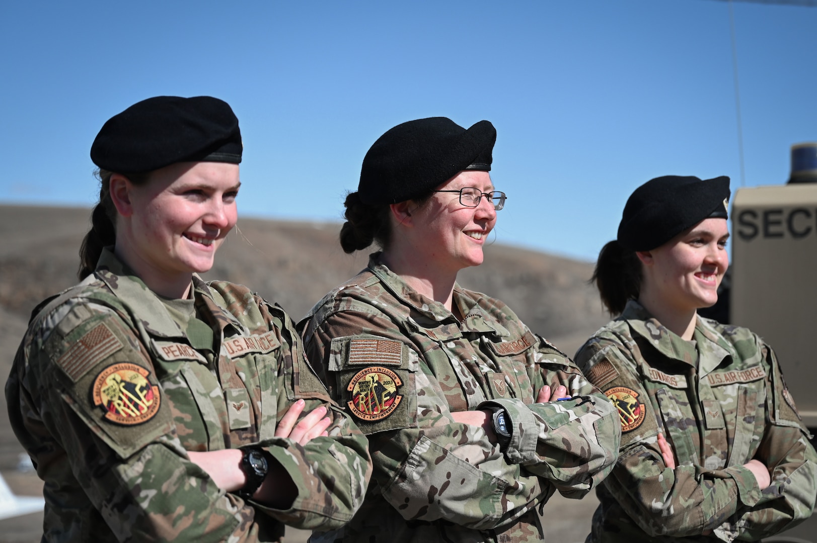Three women in military uniform and black berets stand for a portrait against blue sky.