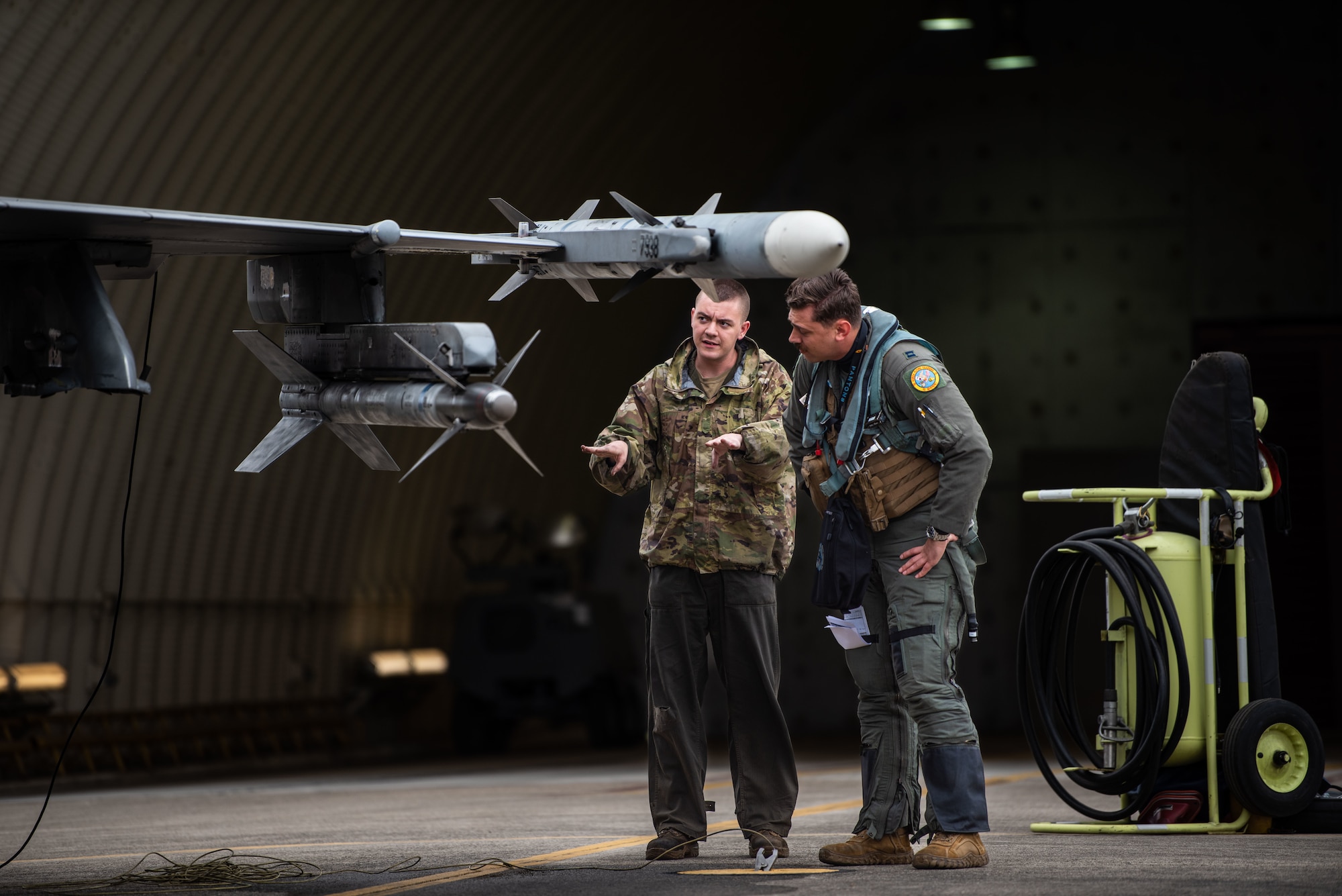 A maintainer talks with his hands during a pre-flight inspection with a pilot who is looking at a munition