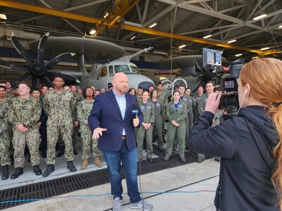 Sailors stand behind a newscaster in their hangar.