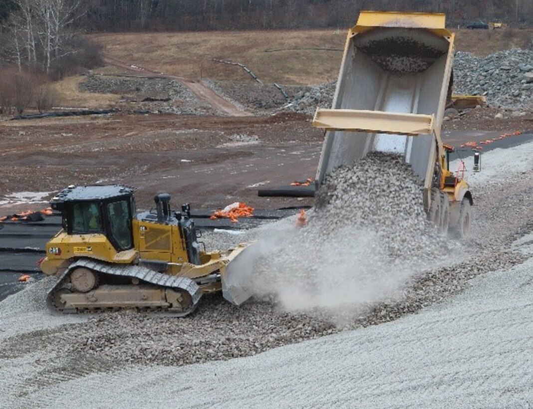 A dump truck deposits aggregate material and a bulldozer pushes it up the slope of a dam