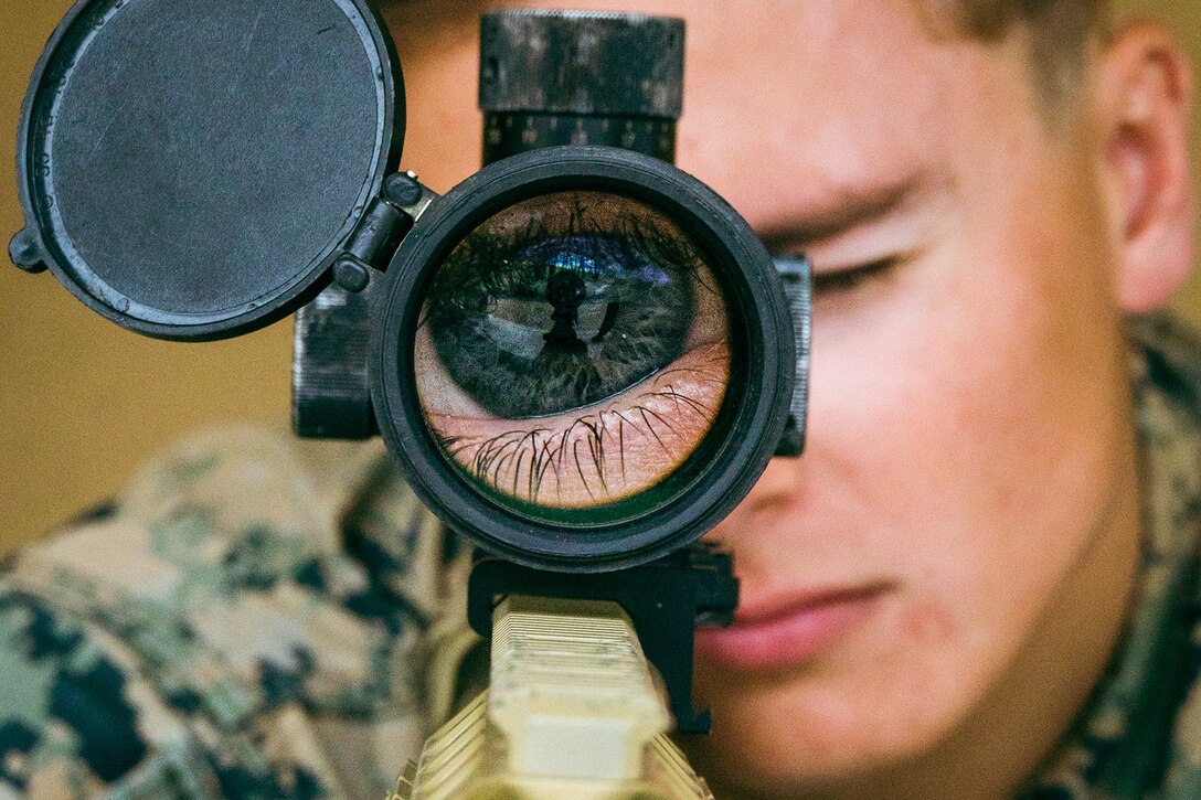 A Marine looks through the scope of a weapon. The Marine’s eye is shown magnified in the scope.