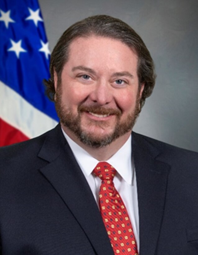 Photo of man in blue blazer, light blue shirt red tie in front of U.S. flag.