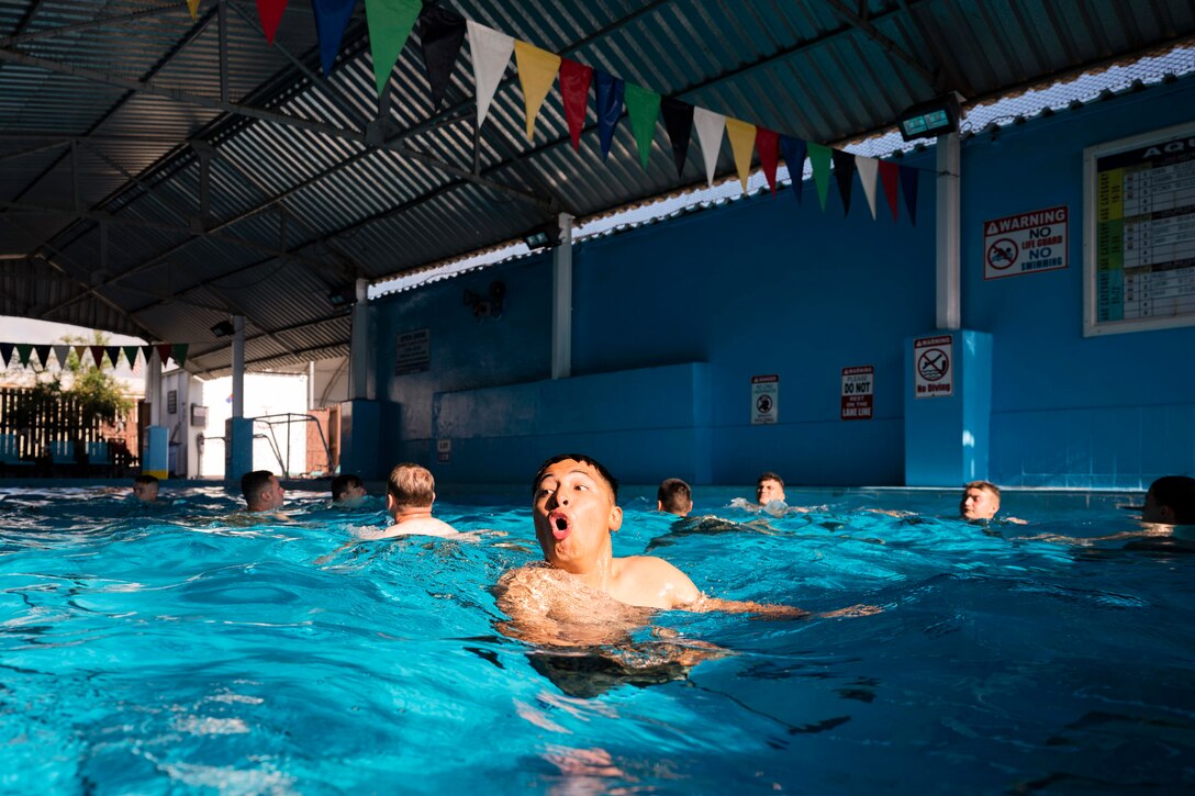 A soldier gasps for air while floating in a pool as fellow soldiers tread water in the background.