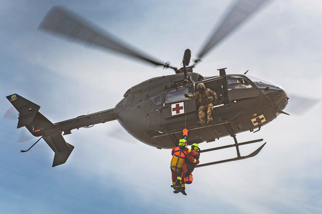 A guardsman sitting in the open doorway of an airborne helicopter watches as two firefighters connected to a rope are being raised.
