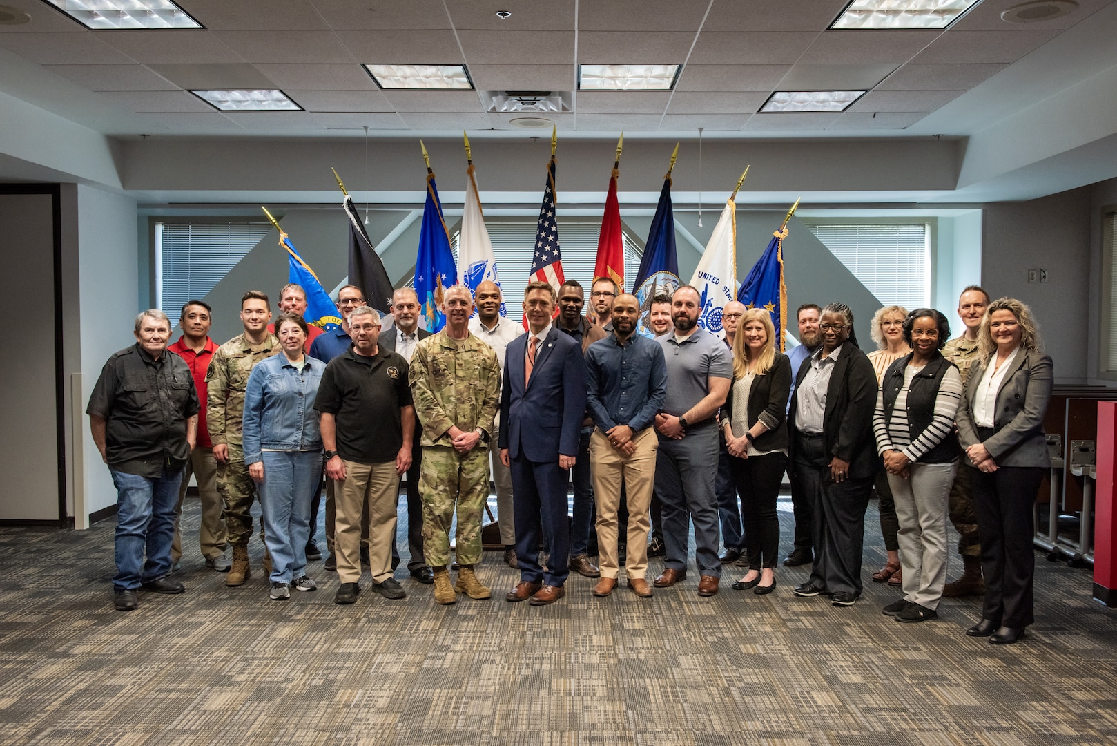 Twenty-four smiling people with a mix of military and civilian. The military are wearing camoflage OCPs and the civilians are wearing a varying combination of business casual and casual.  There are men and women in the photo. They are standing in front of an array of military flags inside a conference room with white walls and grey carpet.