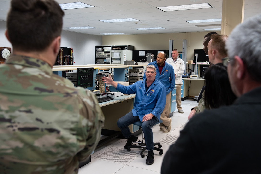 A man in blue lab coat is sitting in a chair with his out gesturing toward equipment. Two men stand behind him - one with a blue lab coat and another with a white lab coat.