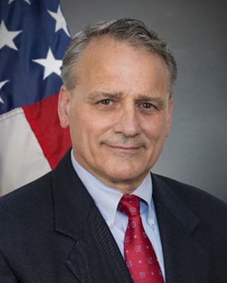 Photo of man in navy blazer, light blue shirt with red tie in front of U.S. flag