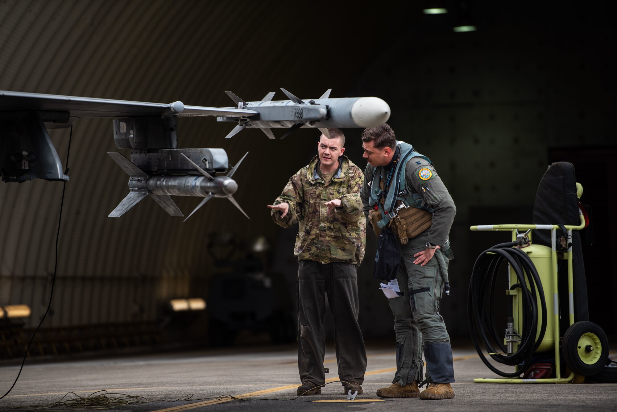 A maintainer talks with his hands during a pre-flight inspection with a pilot who is looking at a munition