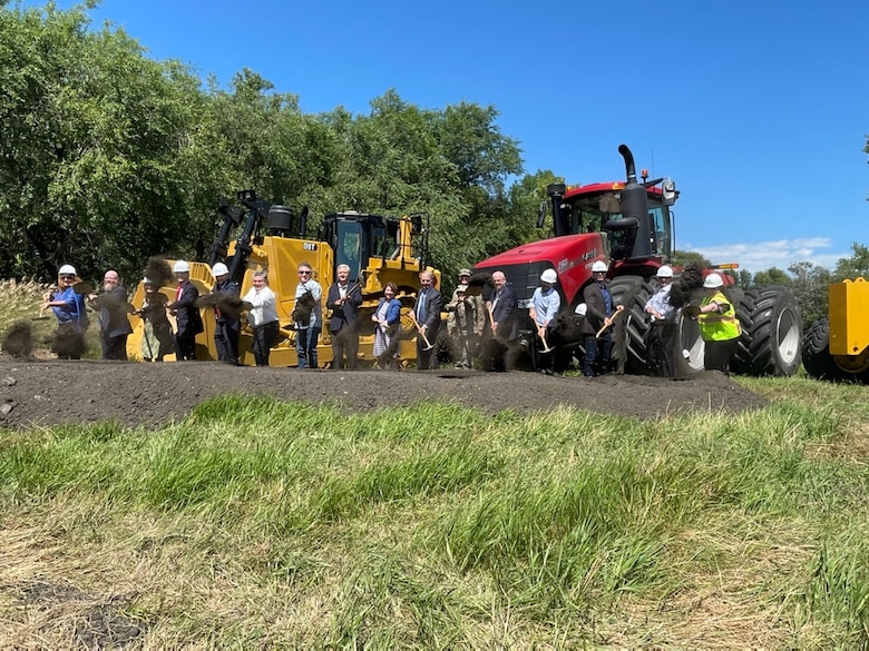 A group of people shovel dirt in front of construction equipment.