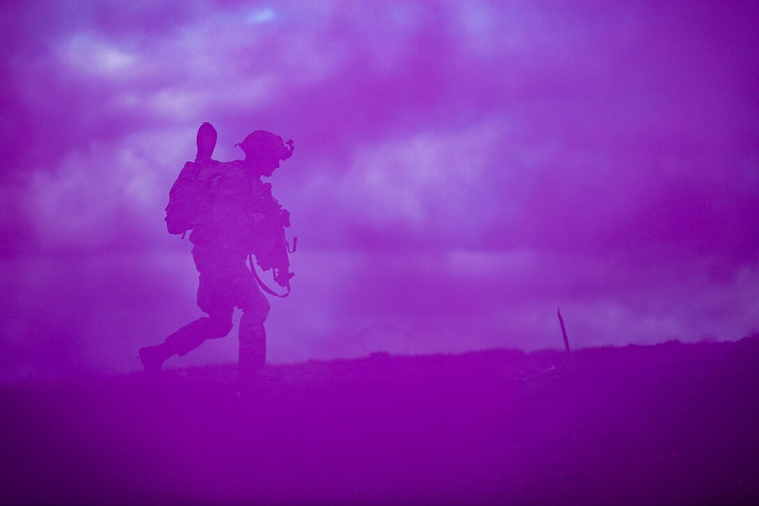 A soldier carrying a weapon and a ruck sack walks through a purple haze of smoke.