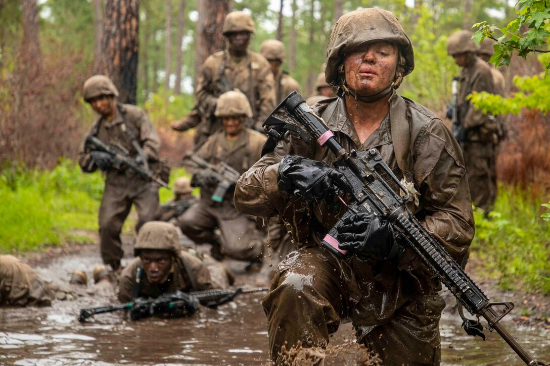 Recruits move through shallow muddy water while holding weapons.
