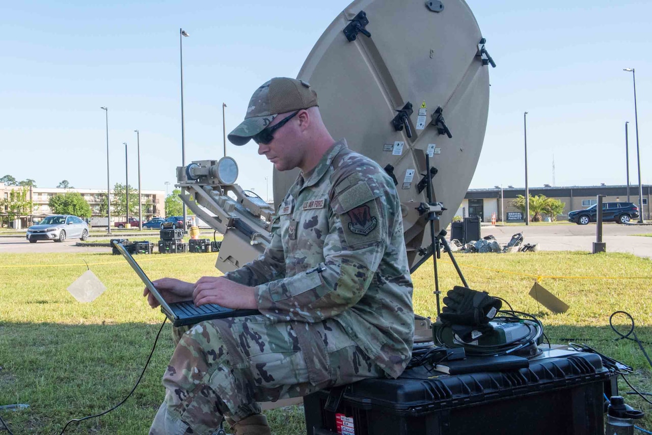 An airman sits on a storage container as he works on a laptop.