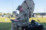 An airman sits on a storage container as he works on a laptop.