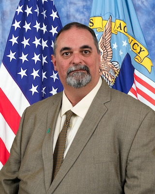 Official photo of Richard TeBeau in brown suit with American and DLA flags in the background