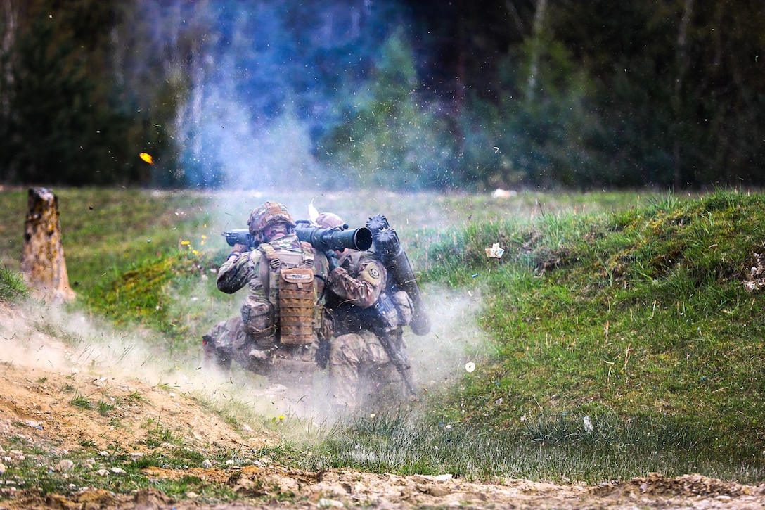 Two soldiers kneel while holding equipment to participate in a live fire exercise.