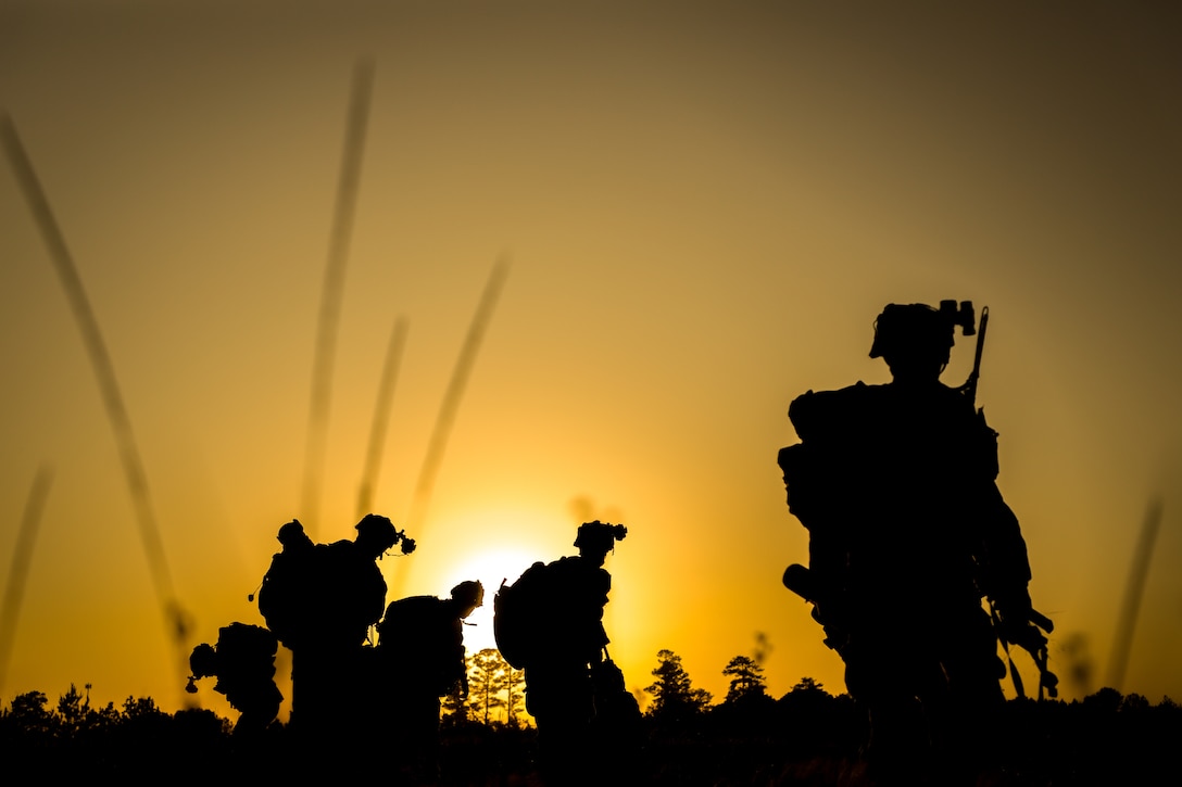 Five service members in silhouette wearing headgear search a field amid a sunny orange backdrop.