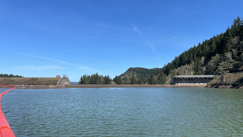 A hill slopes in to the picture from the right, meeting the spillway of a dam. The top of the photo is blue sky, and the bottom of the photo is the water behind the dam, which makes up Cottage Grove reservoir. On the bottom right, a bright orange boom curves across the top of the water toward the dam.