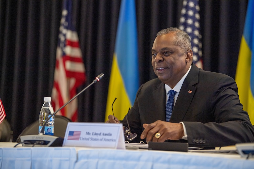 A man in a suit speaks while seated at a table behind a microphone.