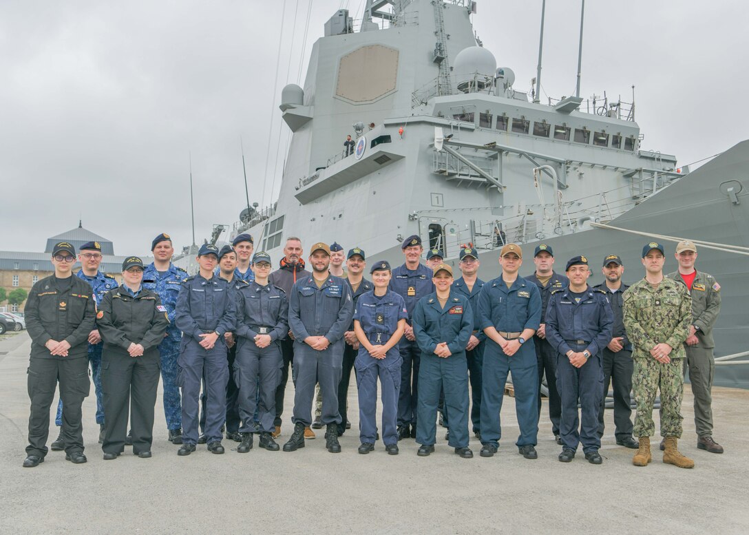 Multinational service members stand for a photo on a pier in front of a ship.