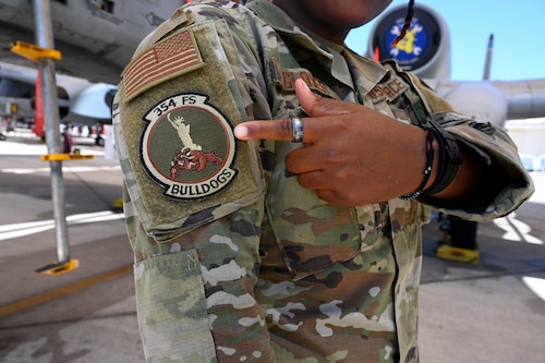 Senior Airman Gabrielle Glover, 354th Fighter Squadron all-source intelligence analyst, points to her squadron patch at Davis-Monthan Air Force Base, Ariz., April 25, 2023. At the 354th FS, Glover equips the pilots with real-life information to replicate the battle spaces that the pilots likely encounter to train for their upcoming missions, on behalf of Air Combat Command. (U.S. Air Force photo by Staff Sgt. Abbey Rieves)