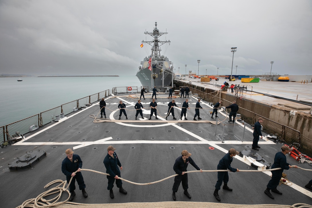Sailors pull ropes preparing to depart a naval station.