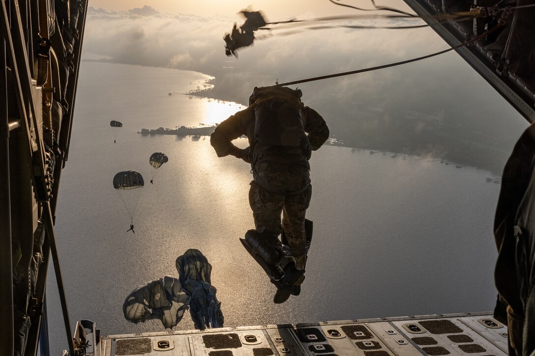 An airman parachutes out of a military aircraft as others descend over water.