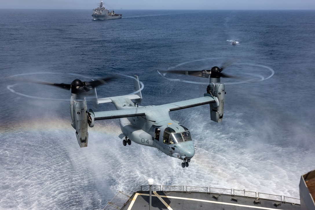 A Marine Corps aircraft lands on a Navy ship as another ship sails in the distance.