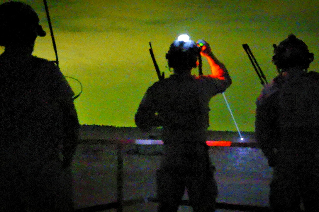 Three airmen shown in silhouette as one shines a spotlight.