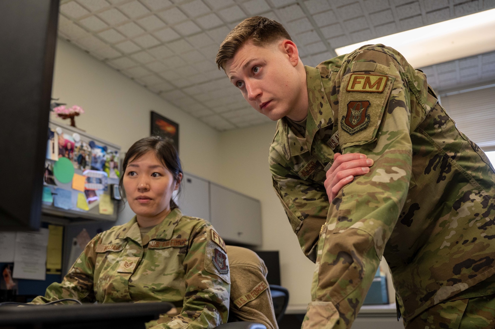 A seated female Airman, left, receives instruction from a standing male Airman, right, as he points at a computer monitor