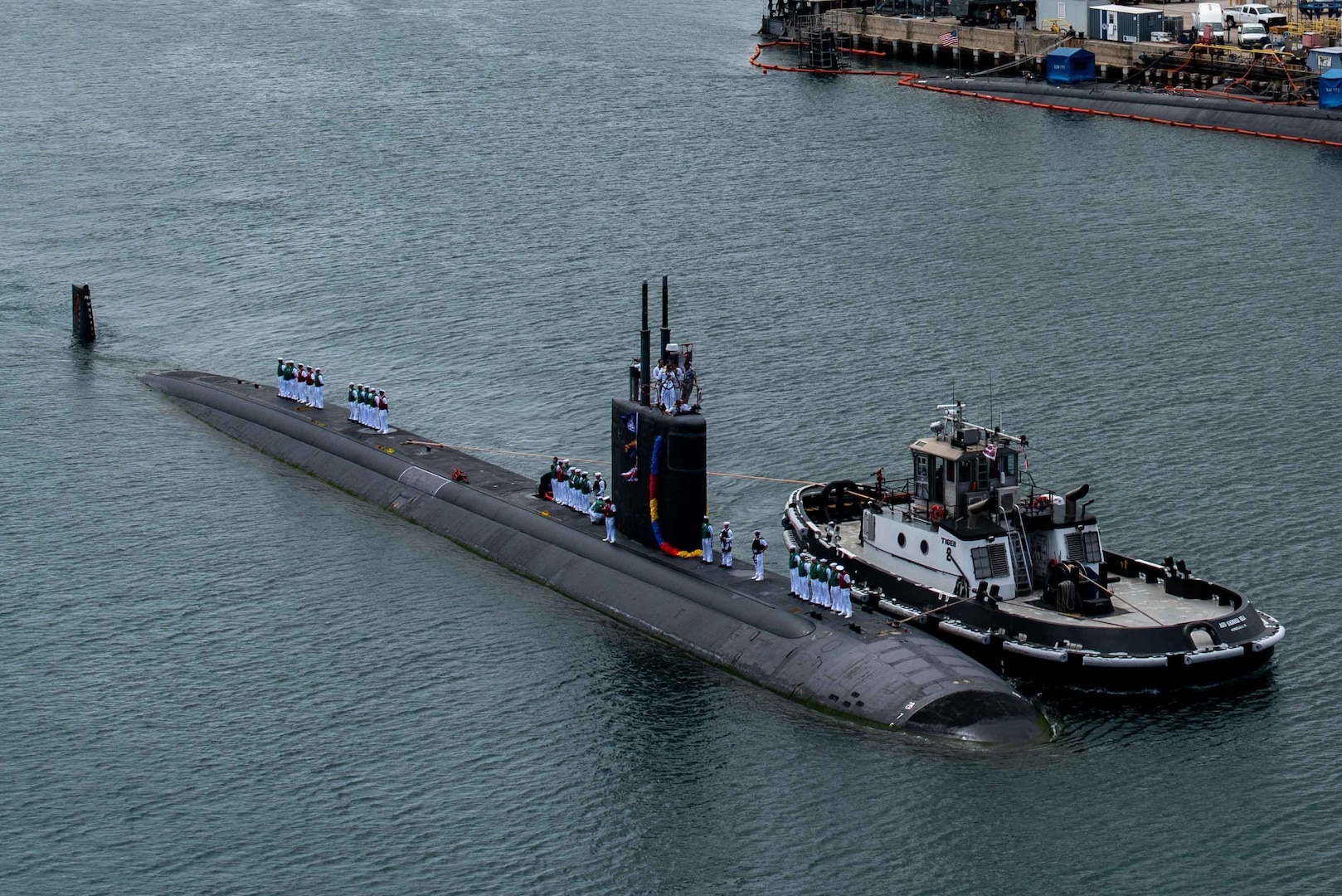 An overhead view of a surfaced submarine moving into a port facility.