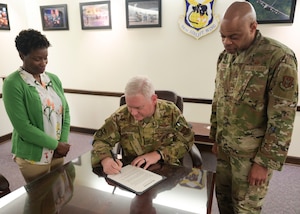 Woman in green sweater stands next to two men in Air Force uniform while one man sitting, signs a mental health awareness month proclamation.
