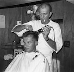 "Haircut -- L. N. Baker, EN3, USCG getting a haircut from BOSN (W-1) Bernard C. Webber, USCG, acting as barber.  Both are on staff [with USCG] Div. 12." Photo by LCDR R. J. Knapp, USCG, dated 09-24-65.