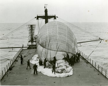 Coast Guardsmen are removing the restraining net from the balloon prior to sending it aloft. The fins are not yet inflated, as they will automatically fill with air as the balloon rises, through a wind tunnel which runs the length of the balloon from the nose to the fins. This photo series was made on March 7-8, 1952, during a test cruise of the USCGC COURIER (WAGR-410),