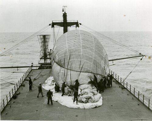 Coast Guardsmen are removing the restraining net from the balloon prior to sending it aloft. The fins are not yet inflated, as they will automatically fill with air as the balloon rises, through a wind tunnel which runs the length of the balloon from the nose to the fins. This photo series was made on March 7-8, 1952, during a test cruise of the USCGC COURIER (WAGR-410),