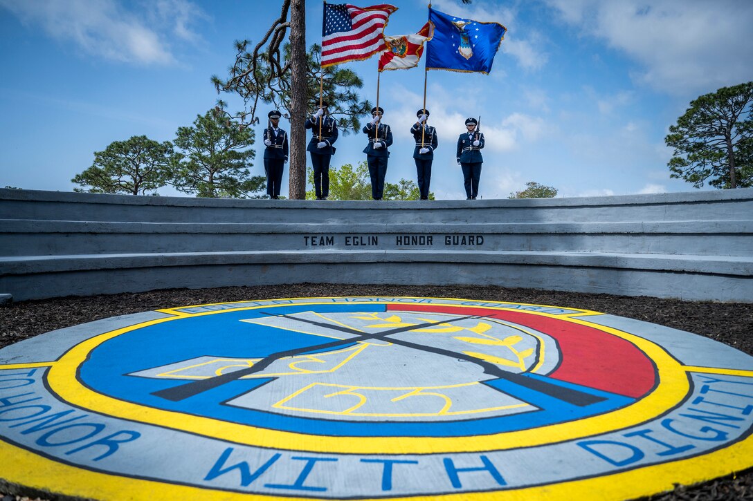 Five airmen in formal uniforms hold flags as they stand at the top of curved steps.