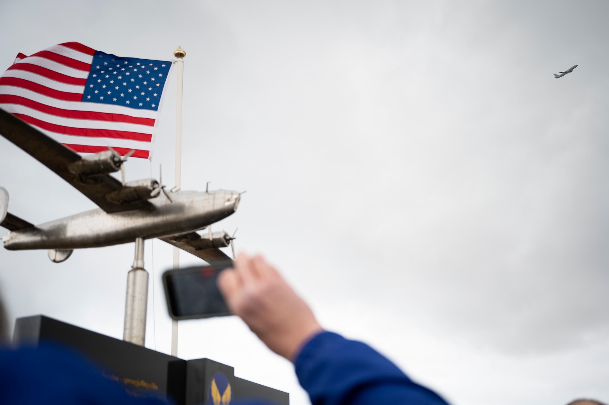 A person takes a photo with their cellphone as a plan flies over a monument.