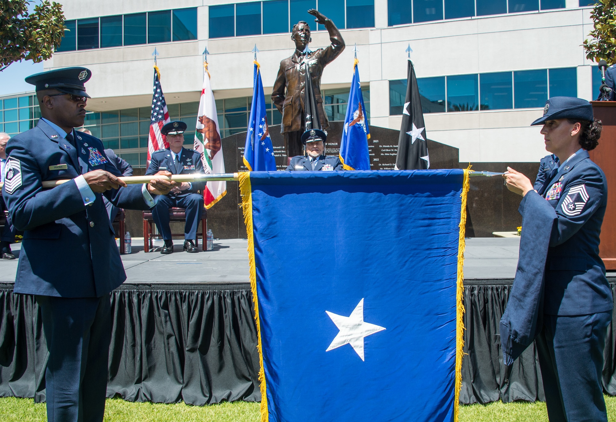 Space Force Chief Master Sgt. Willie Frazier (left) and Air Force Chief Master Sgt. Sarah Morgan unfurl the brigadier general officer’s flag at the Space Systems Command courtyard. The ceremony was held for astronaut and fighter pilot retired Brig. Gen. Buzz Aldrin, who recently received an honorary promotion during a ceremony at Los Angeles Air Force Base, Calif., May 5, 2023. (U.S. Space Force photo by Van Ha)