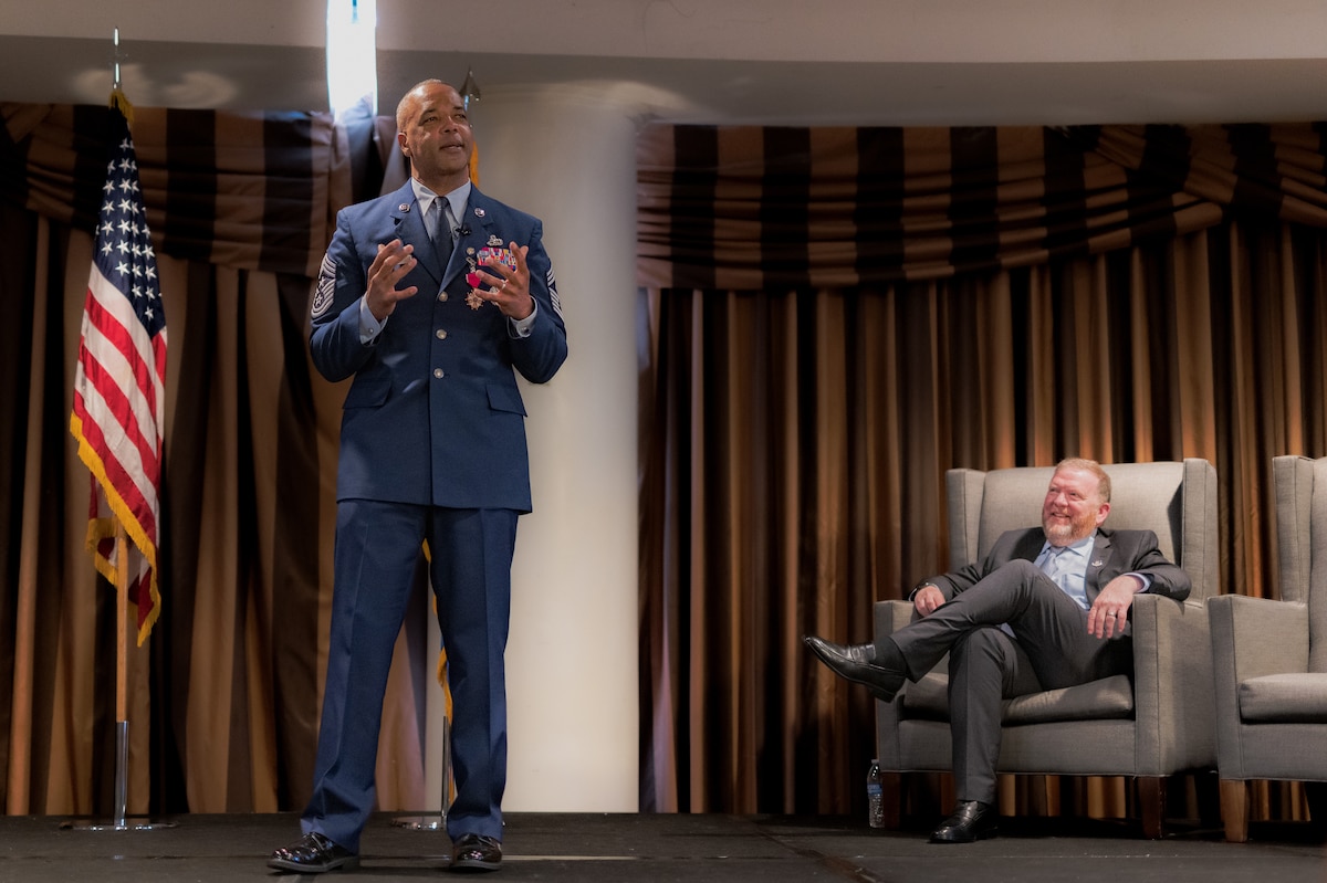 Photo of Chief Master Sgt. Timothy White standing and speaking and Lt. Gen. (Ret.) Richard Scobee sitting and smiling.