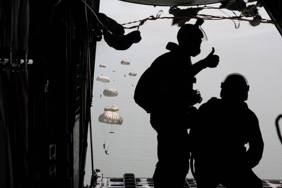 Sailors stand at the back of a military aircraft as others parachute in the background.