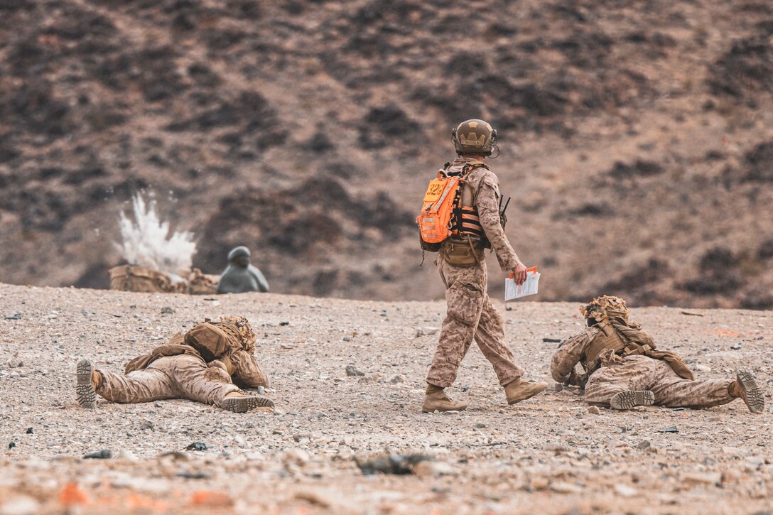 Two Marines fire weapons while laying on the dirt as a fellow Marine observes with a mountain in the background.