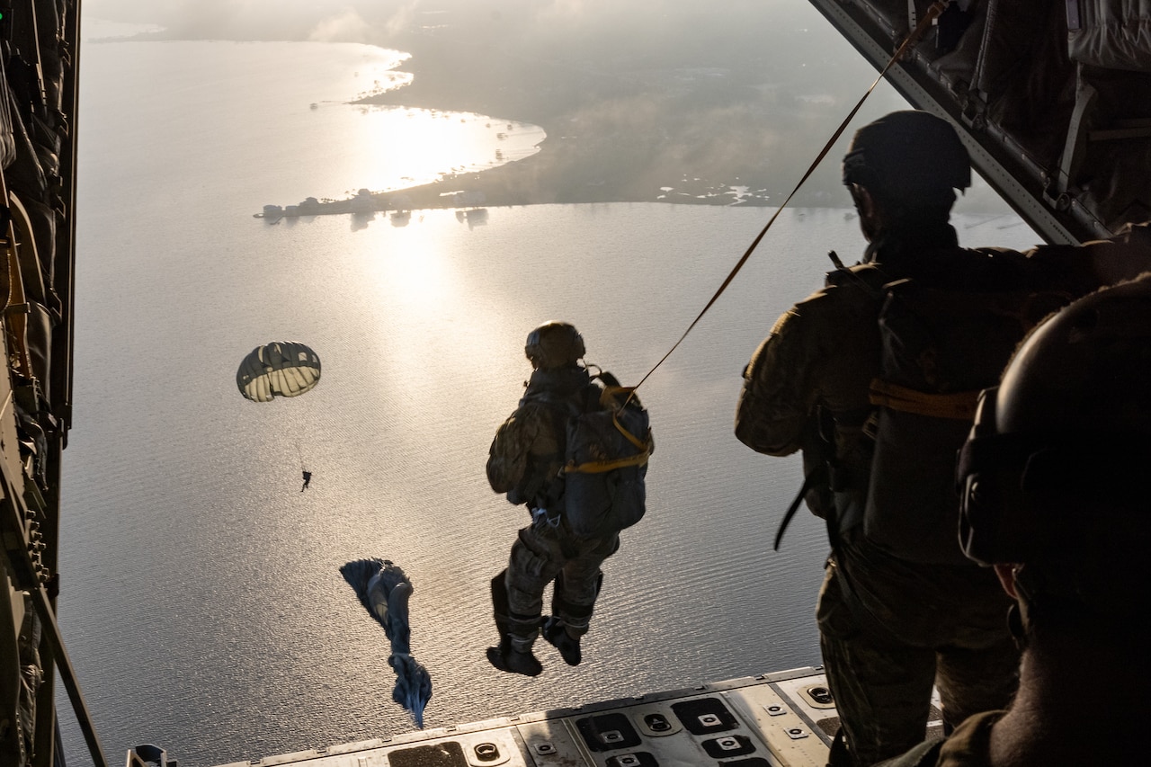 Airmen parachute out of a military aircraft as others descend over water.