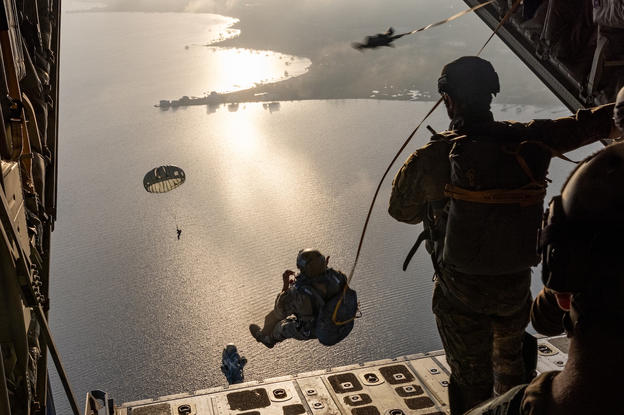 Airmen parachute out of a military aircraft as others descend over water.