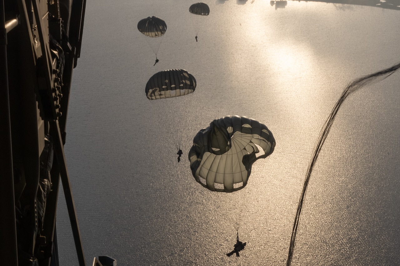 Airmen parachute out of a military aircraft as others descend over water.